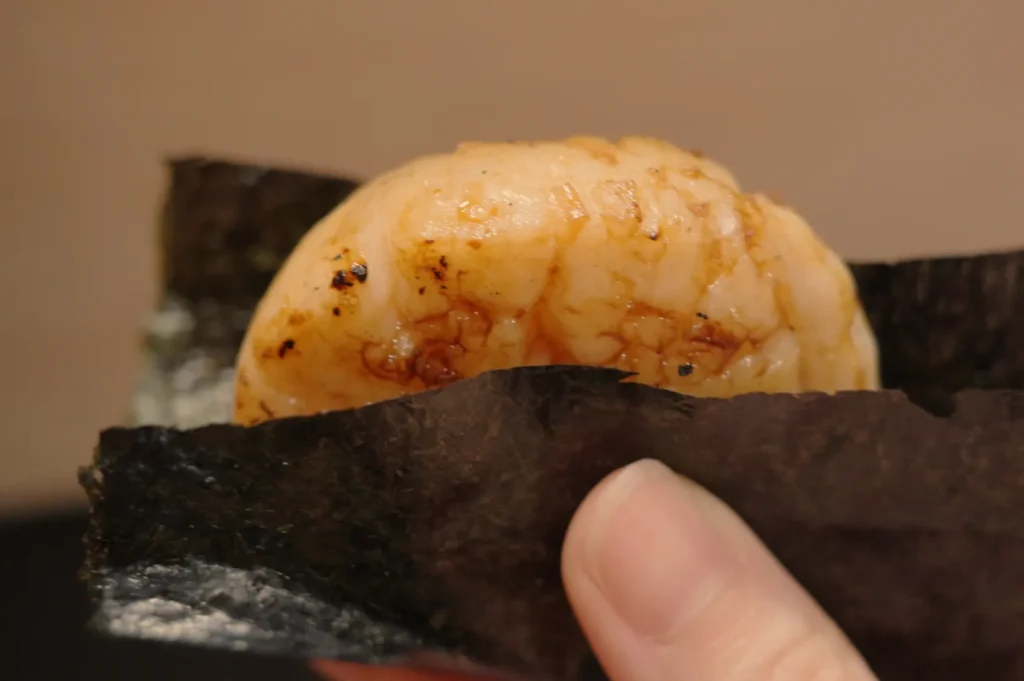 A close-up of a piece of sushi, featuring a piece of seafood on top of a nori seaweed wrap, being held by a hand.