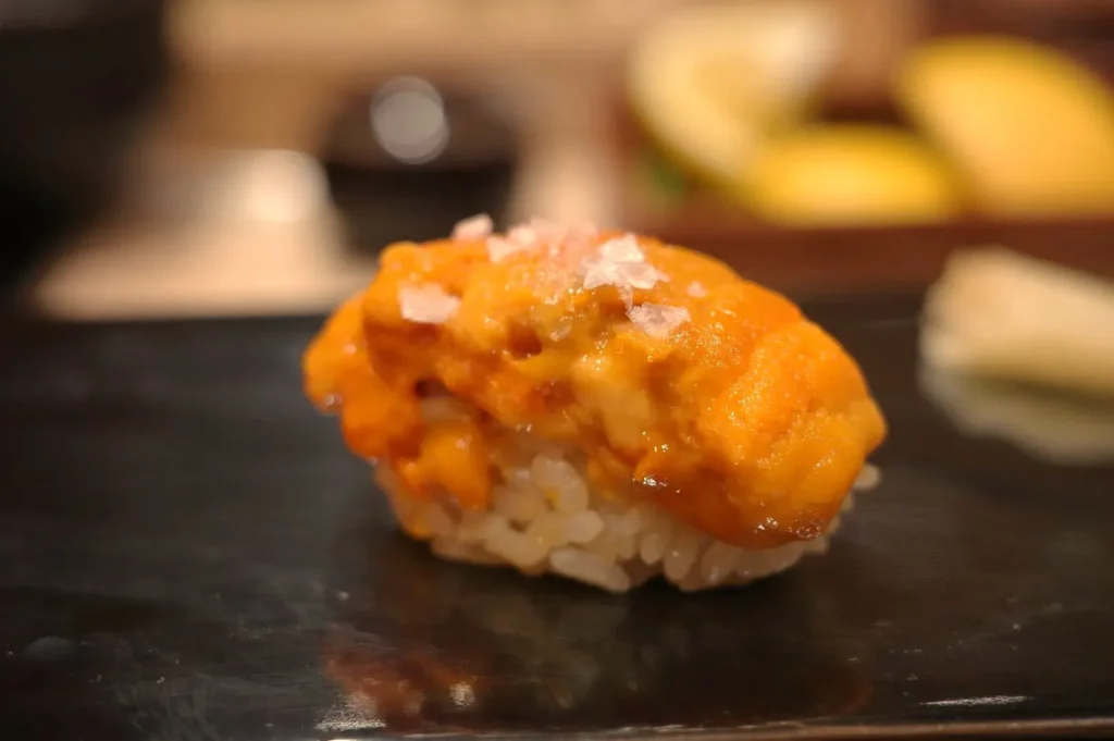 A piece of sushi with a yellow-orange topping, likely sea urchin, on a bed of rice, served on a black plate.