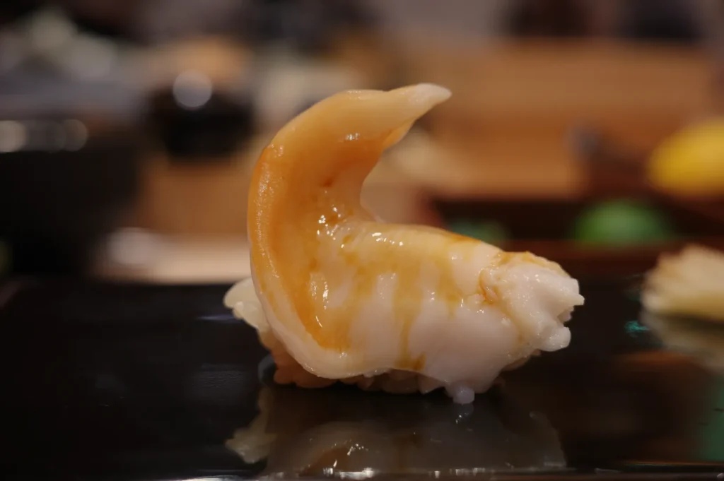 A piece of sushi with a shiny, curled piece of seafood on top, placed on a black plate.