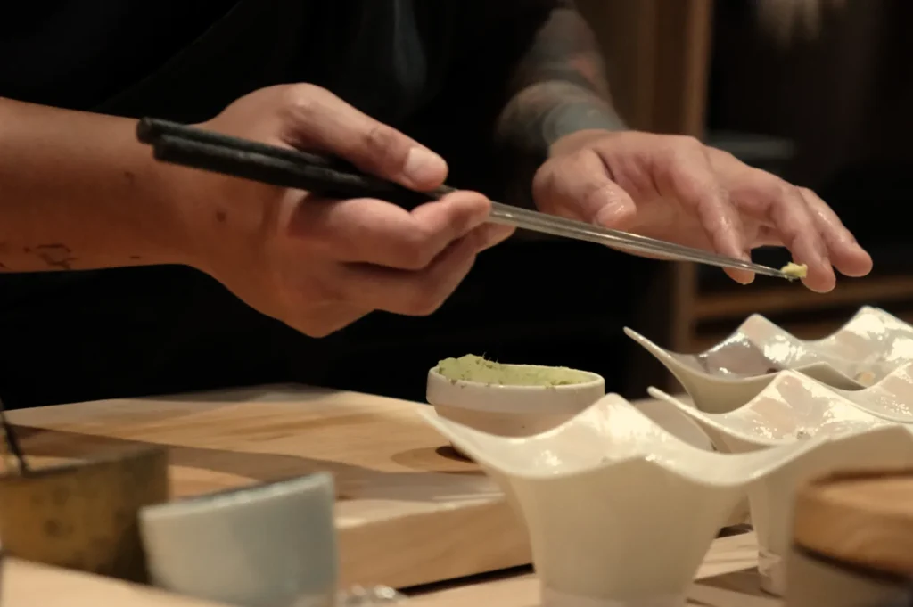 A person using chopsticks to prepare a small portion of green paste, with several small bowls and a wooden platter in the background.