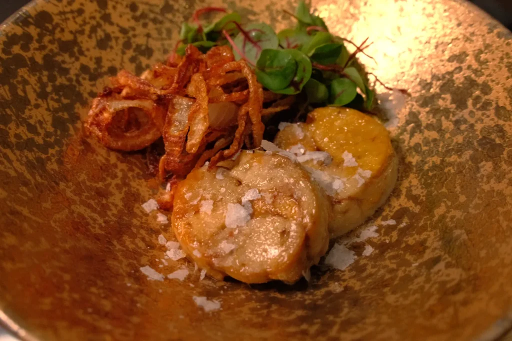 A dish served in a golden bowl containing two round, fried pieces of food, garnished with crispy fried onions and a small salad.