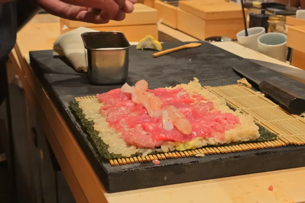 Chef Masa preparing sushi rolls on a bamboo mat with sliced fish and seafood on top of rice and seaweed.