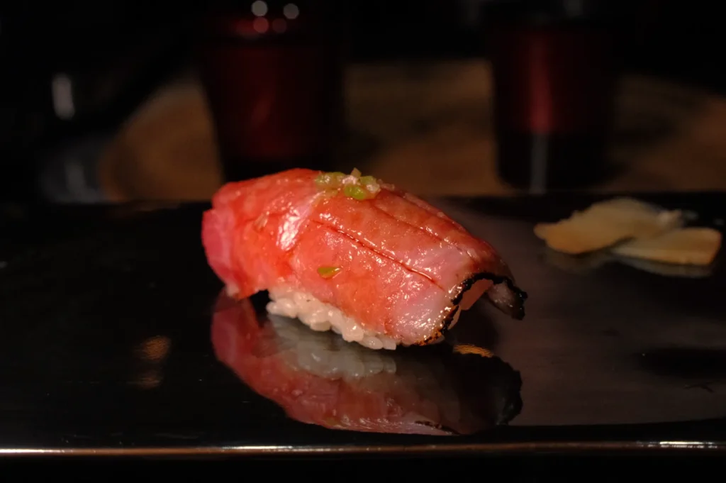 A piece of sushi, specifically nigiri, featuring a slice of fish on top of rice. The fish has a pinkish color and is garnished with a small amount of green onion. The sushi is displayed on a black plate with a hint of ginger in the background.