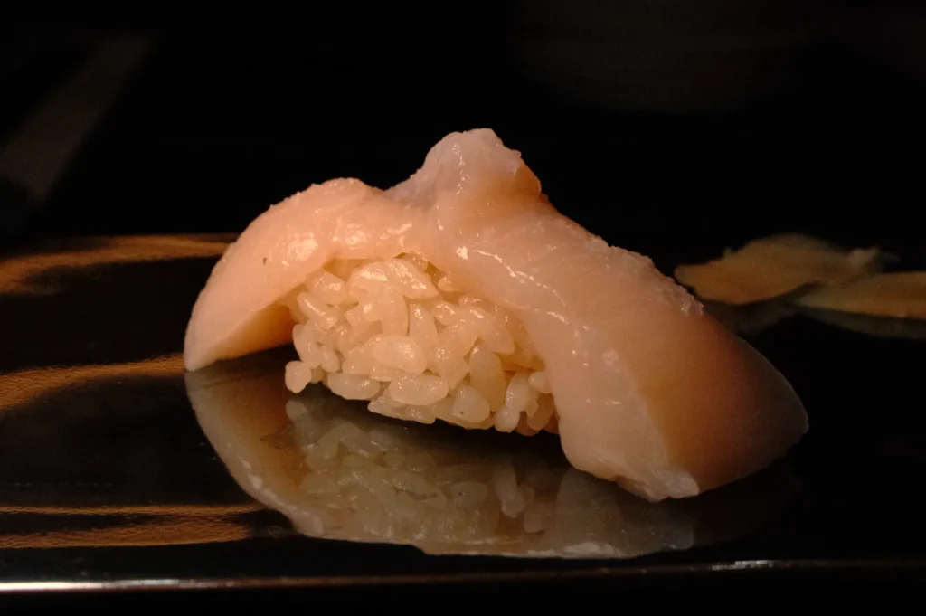 A piece of sushi with a portion of rice topped by a light-colored slice of seafood, reflecting on a dark surface.