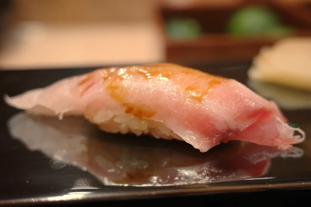 A piece of sushi on a black plate, featuring a slice of fish atop a mound of rice, with a glossy appearance.