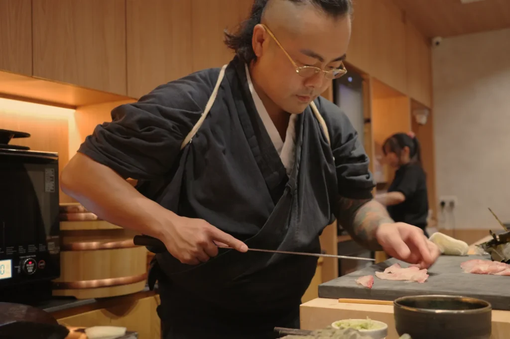 Chef Masa in a traditional attire meticulously prepares sushi in a modern kitchen setting.