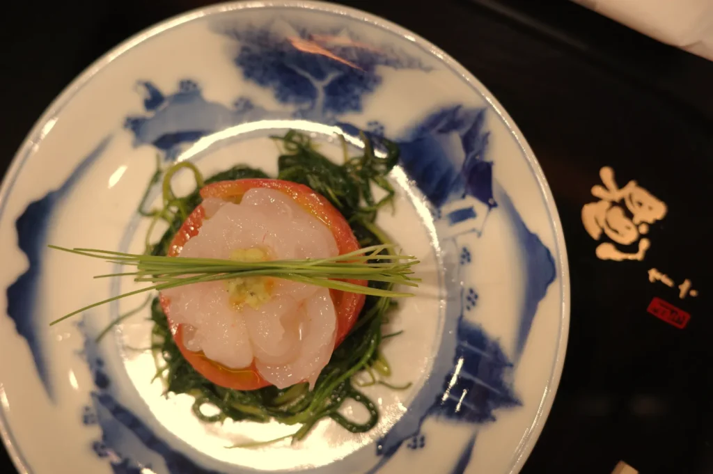 A beautifully plated dish featuring a circular mound of seafood topped with green chives, surrounded by a bed of green seaweed, served on a blue and white patterned plate.