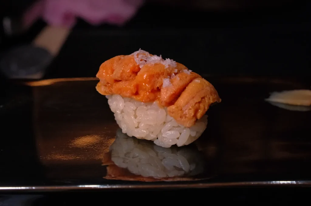 A piece of sushi with orange sea urchin (uni) on top of a small ball of white rice, presented on a dark plate.