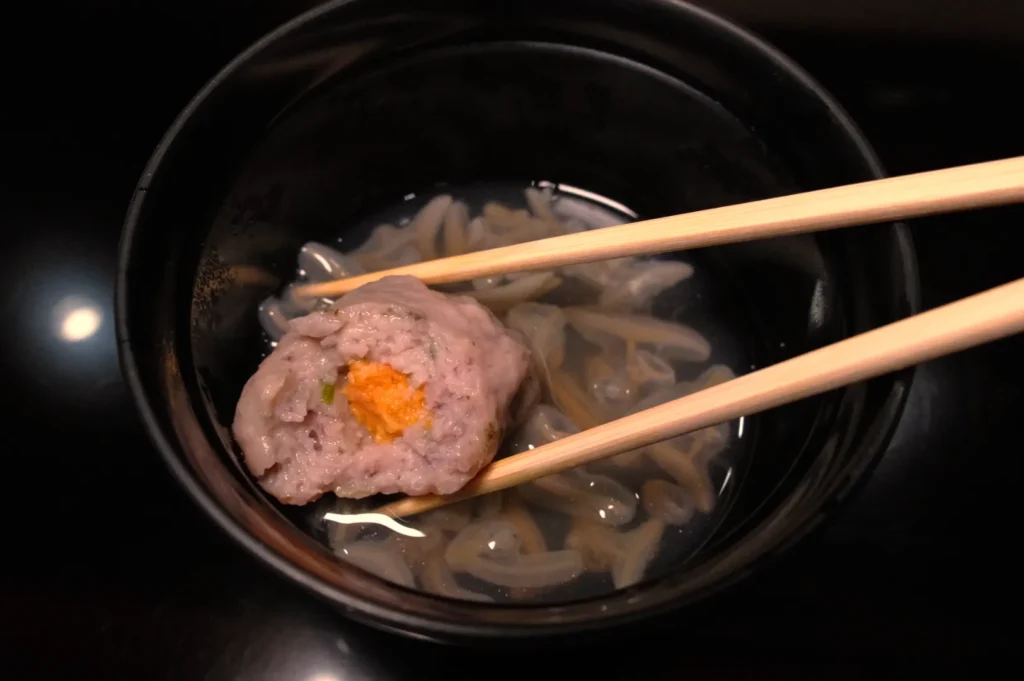 A black bowl containing a clear broth with small noodles and a fish ball that has an orange piece inside, being held by chopsticks.