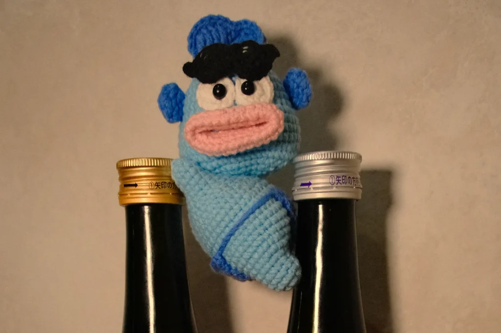 A knitted blue and pink puppet character with black hair, positioned between two bottles.