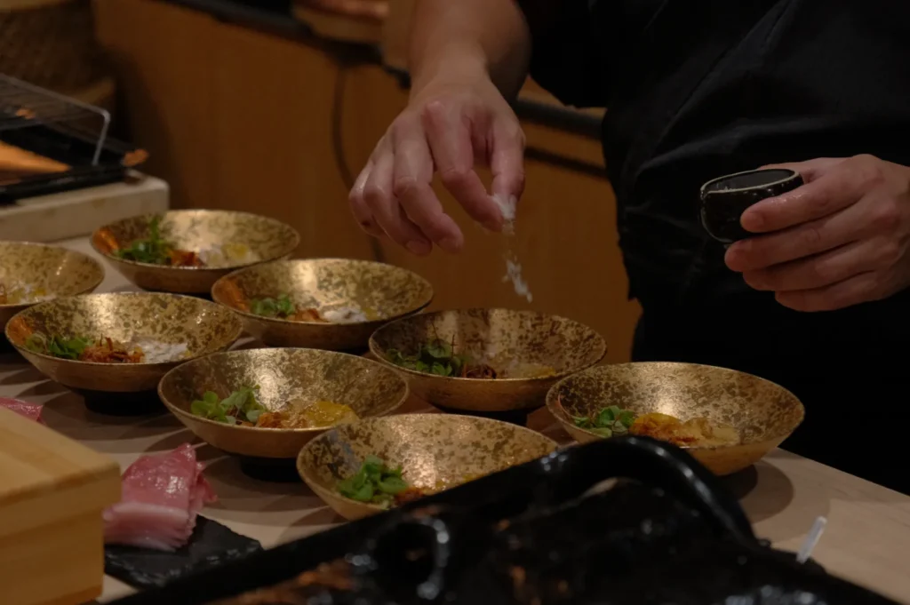 Chef Masa sprinkling seasoning over several decorative bowls filled with food on a countertop.