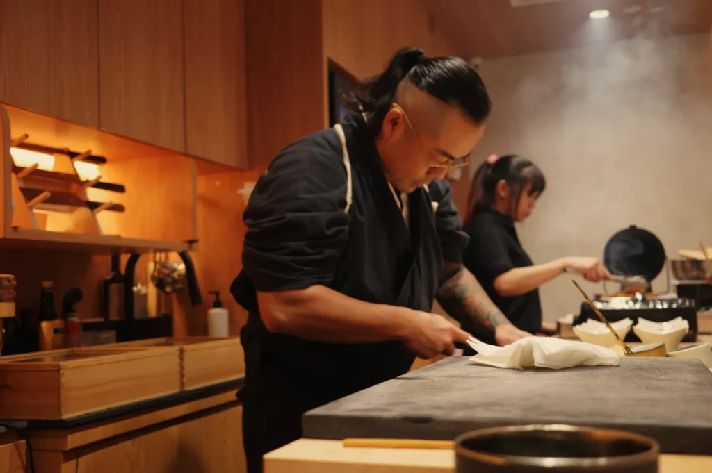 Chef Masa in a dark outfit preparing food in a kitchen, with another person cooking in the background.