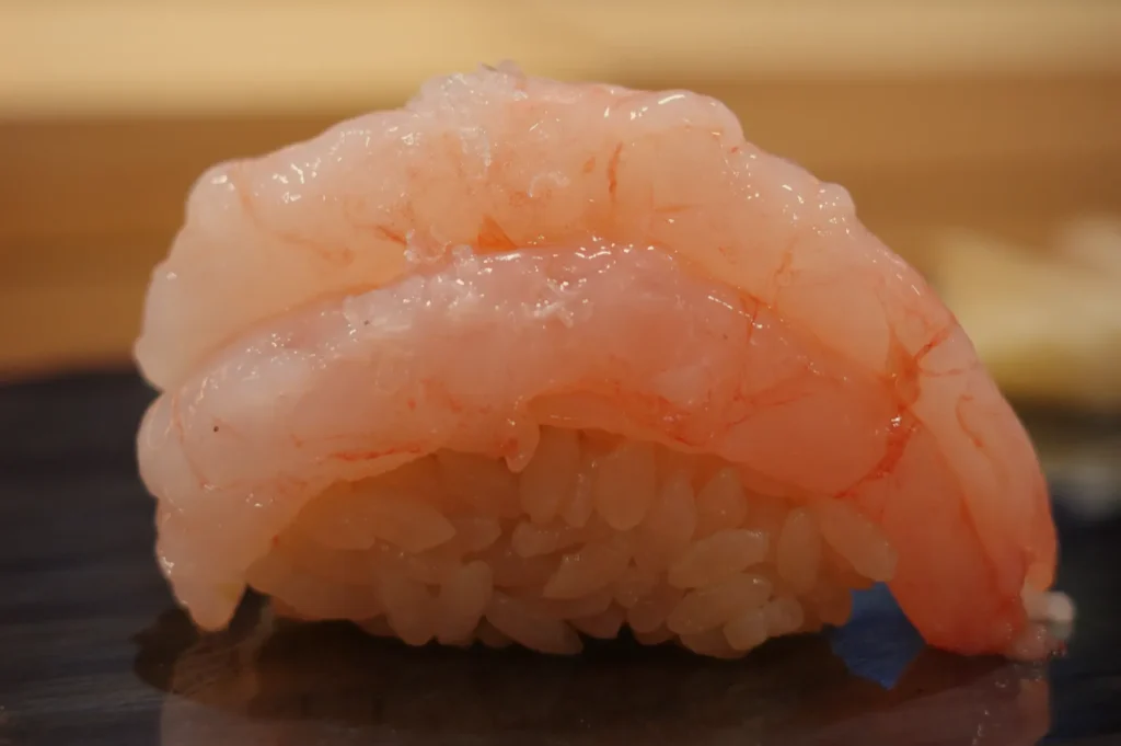 A piece of sushi featuring a translucent pink shrimp topping over a mound of vinegared rice.