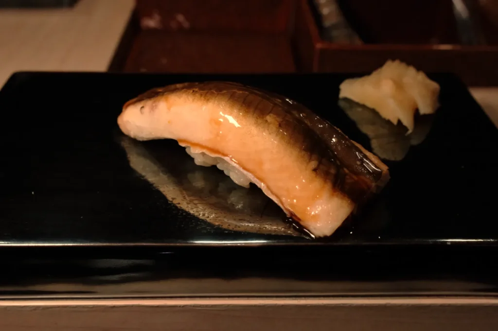 A piece of sushi featuring eel on a black plate, accompanied by a small serving of ginger.