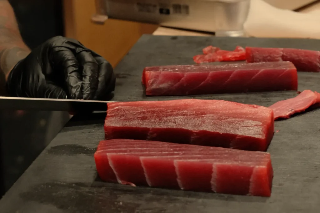 A person with a black glove is slicing raw tuna on a black cutting board. Several pieces of tuna are arranged on the board.