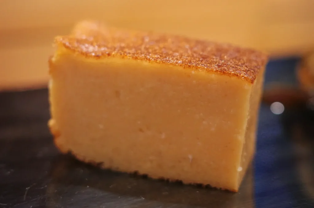 A slice of golden-brown, smooth-textured cake, possibly a type of custard or flan, placed on a dark surface.