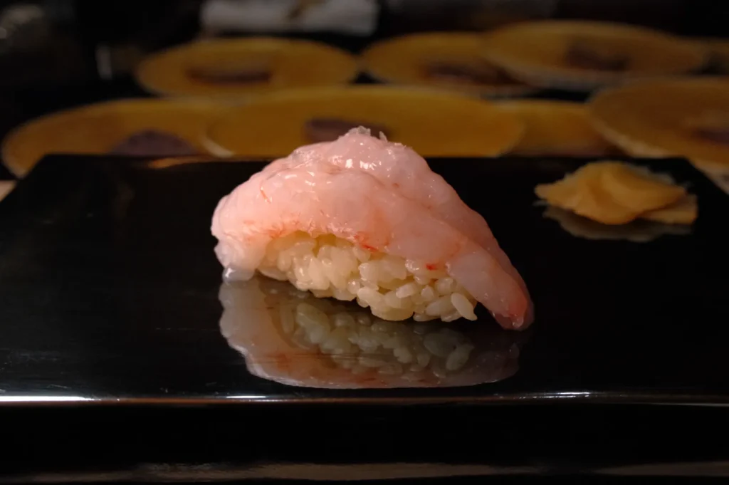 A piece of sushi with shrimp on top, served on a black plate, with some ginger on the side.