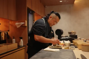 A focused Chef Masa in a black uniform prepares a dish at a wooden restaurant counter, arranging ingredients and tools in a warm, calm kitchen.