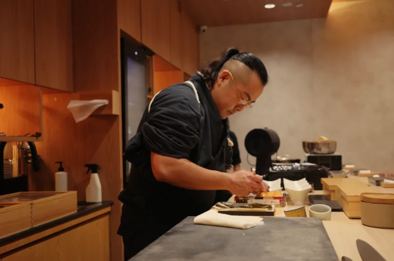 A focused Chef Masa in a black uniform prepares a dish at a wooden restaurant counter, arranging ingredients and tools in a warm, calm kitchen.
