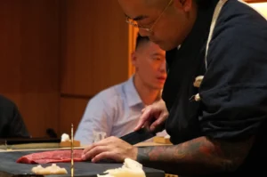 Chef Masa skillfully slicing raw fish on a cutting board in a restaurant setting, with diners in the background.