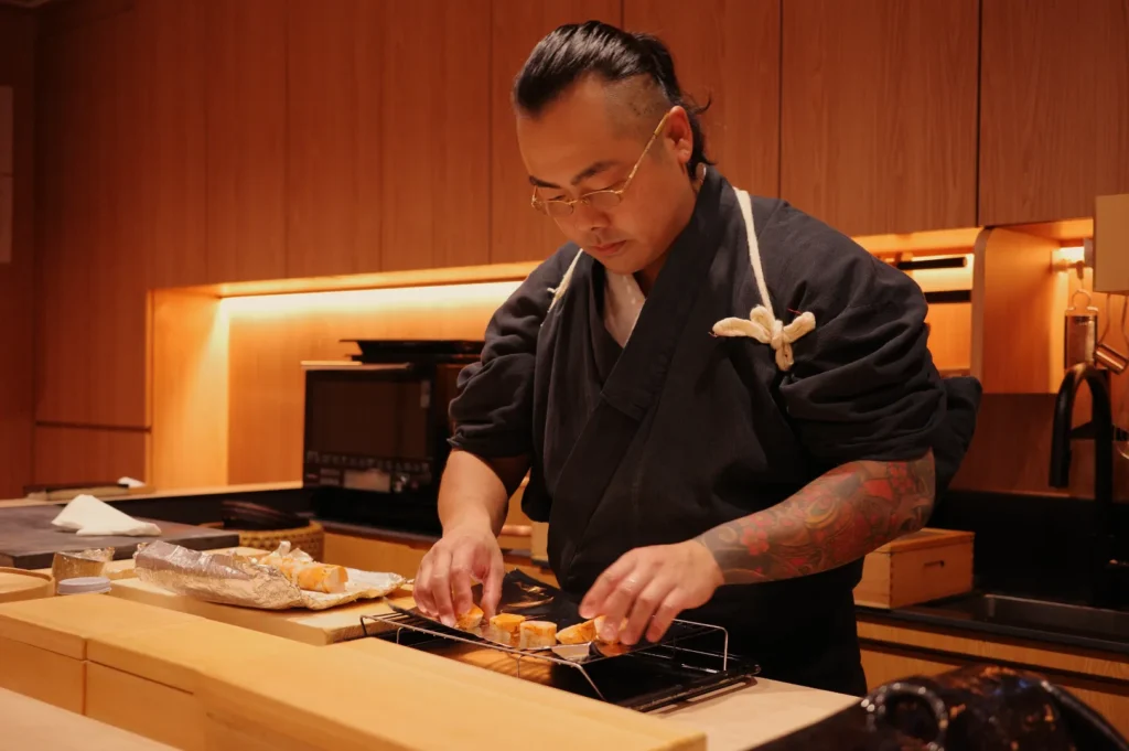 Chef Masa in a black traditional Japanese uniform preparing sushi in a modern kitchen with wooden finishes.