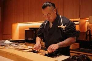 Chef Masa in a black traditional Japanese uniform preparing sushi in a modern kitchen with wooden finishes.