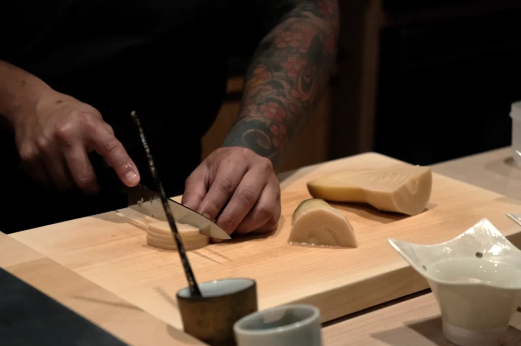 Chef Masa slicing a piece of food on a wooden cutting board, with a blurred background.