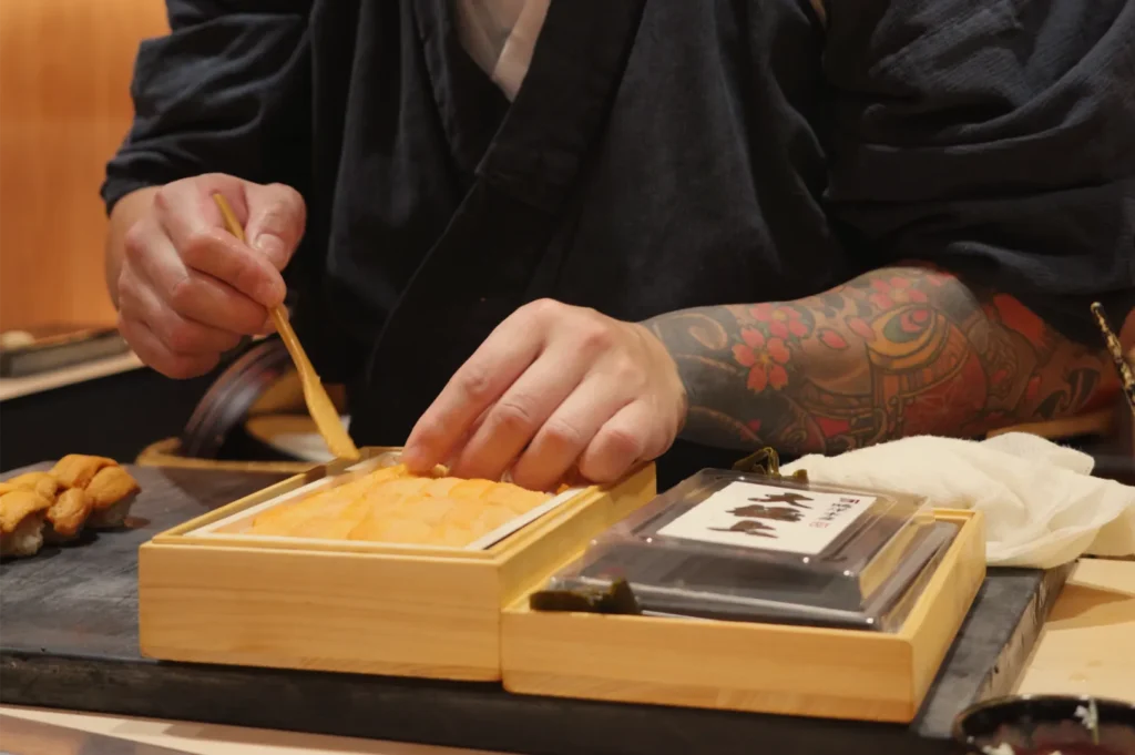 Tattooed chef in black clothing uses a brush to prepare food in a wooden serving box, creating an intimate, artisanal dining scene.