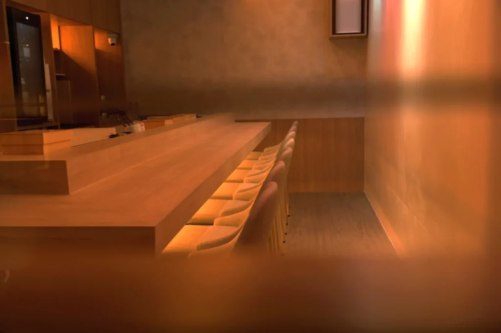 A minimalist interior of a restaurant or bar with a wooden counter and several empty seats, softly lit.