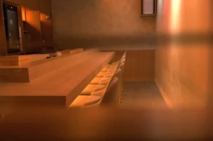 A minimalist interior of a restaurant or bar with a wooden counter and several empty seats, softly lit.