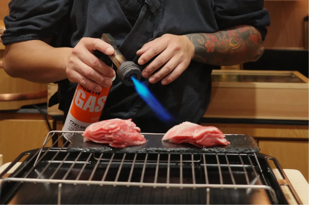 Tattooed cook uses a blue-flame torch and gas canister to sear raw meat on a tabletop grill, creating an intense, hands-on cooking scene.