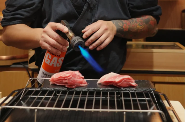 Tattooed cook uses a blue-flame torch and gas canister to sear raw meat on a tabletop grill, creating an intense, hands-on cooking scene.