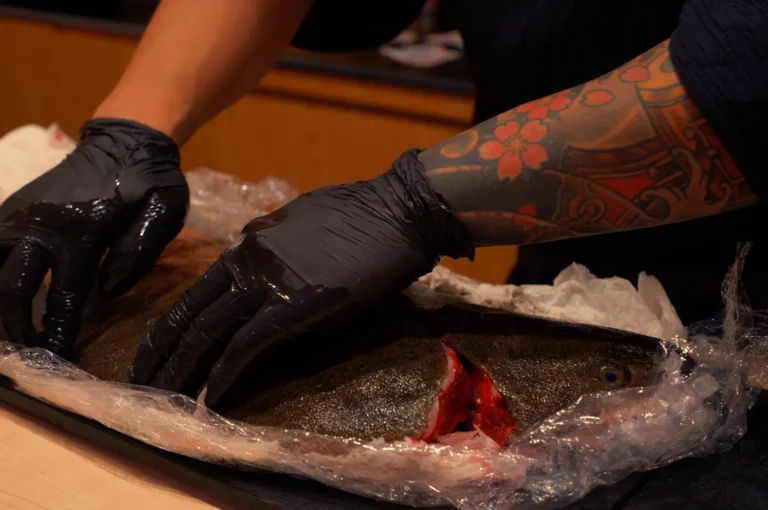 Tattooed arms in black gloves prepare a large fish on ice and plastic wrap, exposing a red cut in a gritty food-preparation scene.