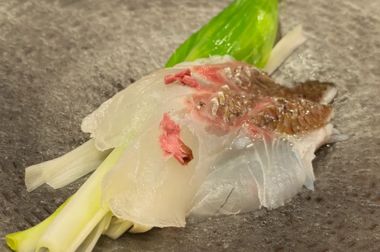 Close-up of translucent sashimi layered with pink petals and brown garnish, served over green shiso and scallions on a dark stone plate.