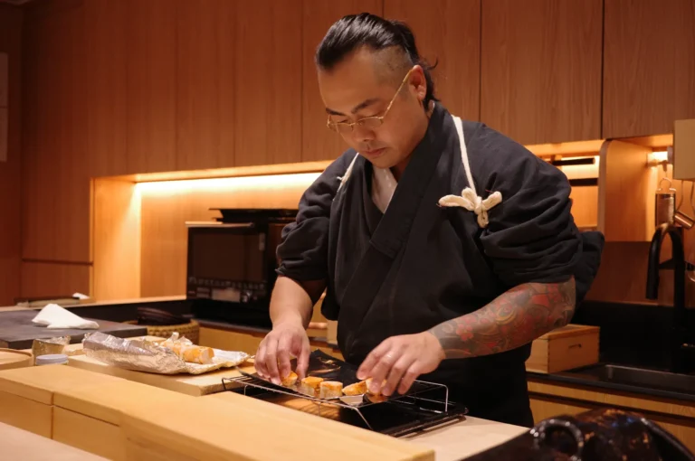 A tattooed sushi chef in a black uniform carefully arranges sushi in a warm, wood-paneled restaurant, creating a focused, intimate atmosphere.