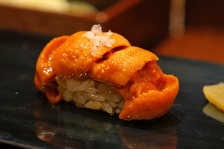 A piece of sushi topped with sea urchin (uni) sits on a black plate, accompanied by a slice of pickled ginger.A piece of sushi featuring orange sea urchin atop a small mound of rice, garnished with a sprinkle of salt, served on a dark plate.