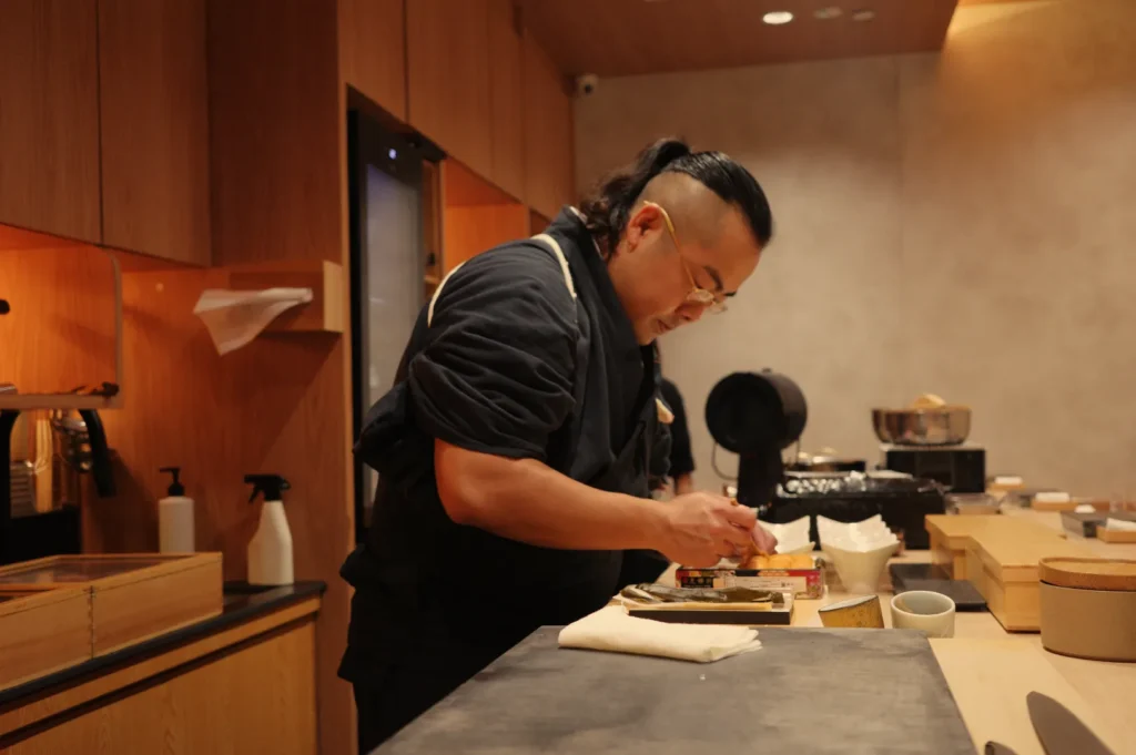 Chef Masa preparing food in a kitchen with wooden walls and various kitchen equipment.