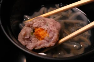 A piece of fish dumpling with orange filling, held by chopsticks above a bowl of soup with thin noodles.