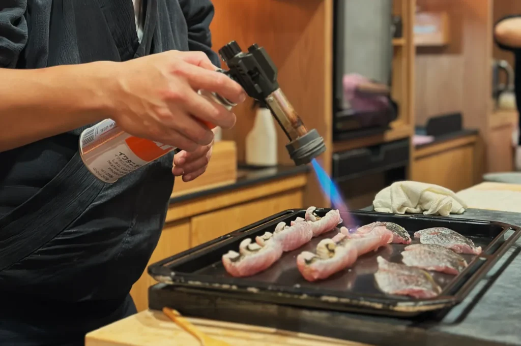 Chef Masa using a culinary torch to sear pieces of fish on a tray in a kitchen.