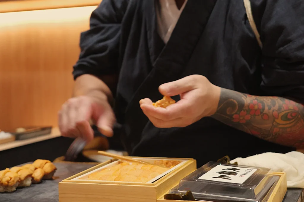 Chef Masa's hand holding a piece of sushi, with a tattooed arm visible and sushi ingredients in wooden boxes nearby.
