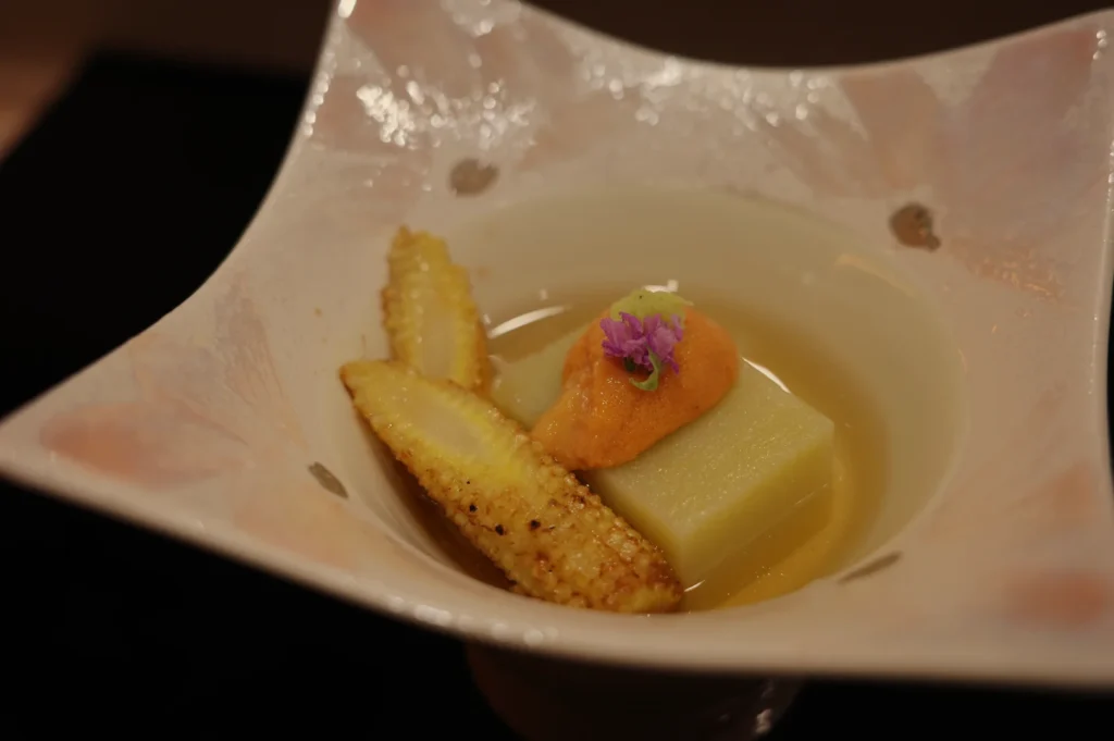A refined plated dish in a pale pink bowl features golden baby corn, a smooth yellow block, and orange purée topped with a purple flower in light sauce.
