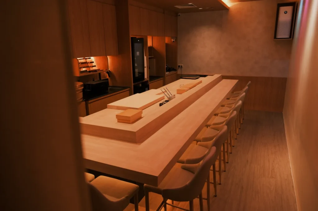 A minimalist kitchen interior with a long wooden table and chairs, featuring modern appliances and warm lighting.