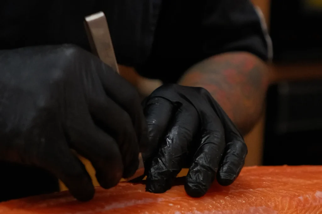 A close-up of Chef Masa’s hands wearing black gloves, skillfully cutting raw salmon with a knife.
