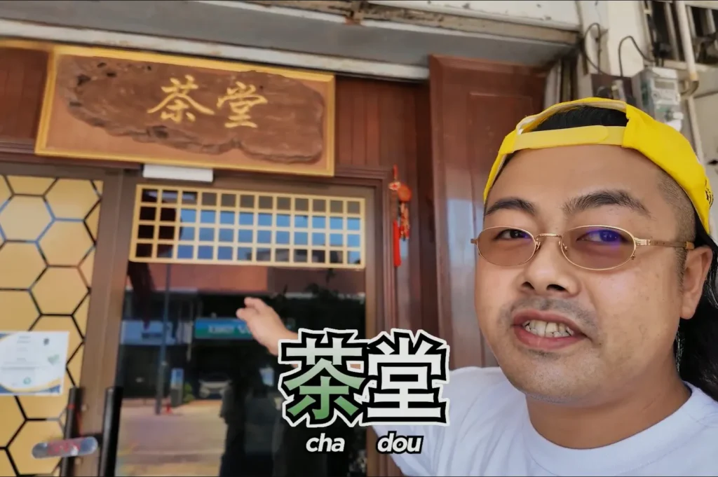 Chef Masa wearing a white shirt and a backwards yellow cap, pointing to a shop with a sign that reads "茶堂" and "cha dou.”