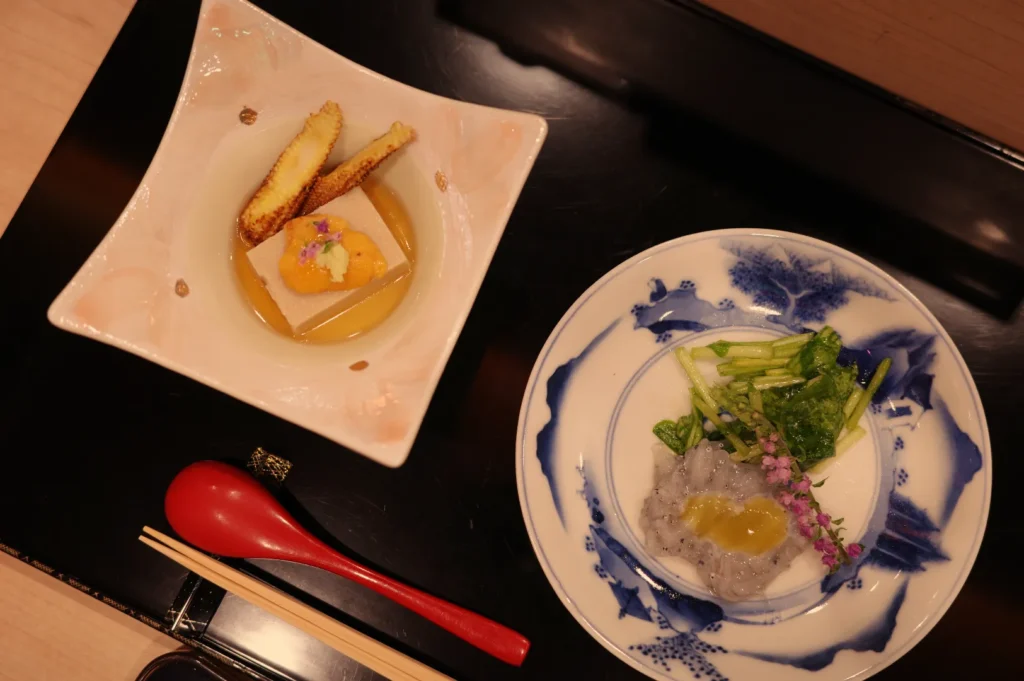 A beautifully arranged Japanese meal featuring a dish with a yellow topping, garnished with flowers, and a side of fried items, accompanied by a plate of rice or a similar dish with vegetables.