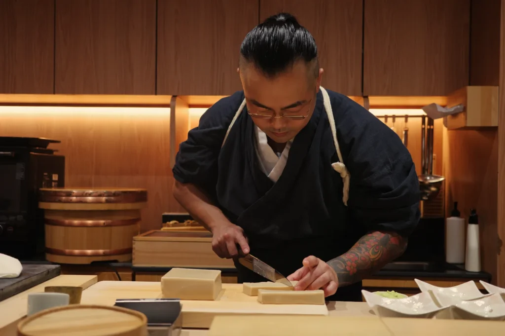 Sushi chef in a dark uniform slices fish at a wooden counter in a warmly lit restaurant, focused, with a tattooed arm and dishes nearby.