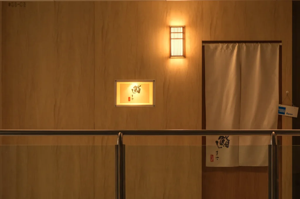 A dimly lit entrance featuring a wooden wall, a small illuminated display case, and a partially closed white curtain with Japanese characters.