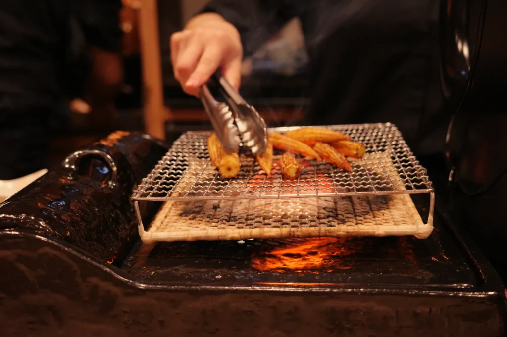 A person using tongs to grill food on a mesh rack over glowing coals.