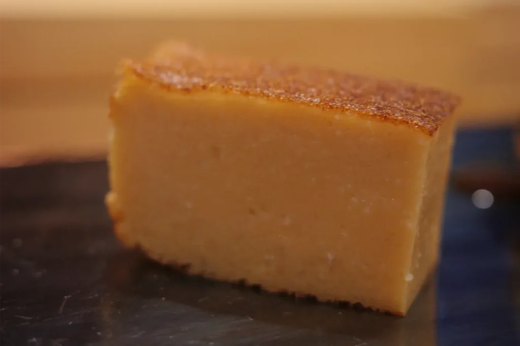 Golden-brown slice of moist sponge cake with a caramelized top, resting on a dark plate against a warm, softly blurred background.