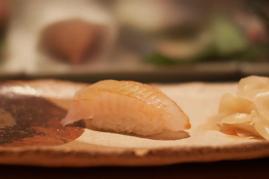 A glossy piece of salmon nigiri sushi rests on a rustic wooden platter beside pale pickled ginger, in warm, softly blurred restaurant light.